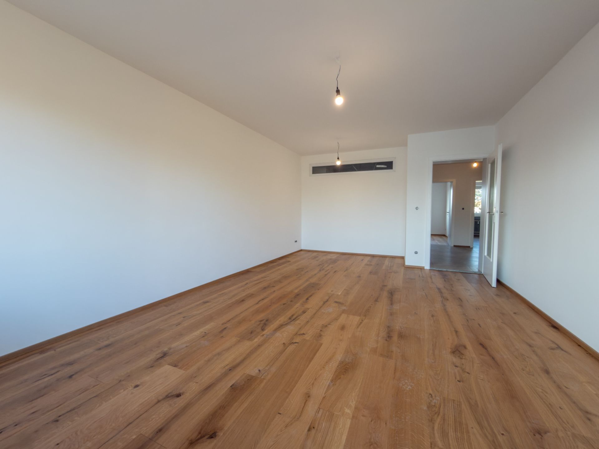 Leeres Zimmer mit Holzboden, weißen Wänden und offener Tür zu weiteren Räumen.