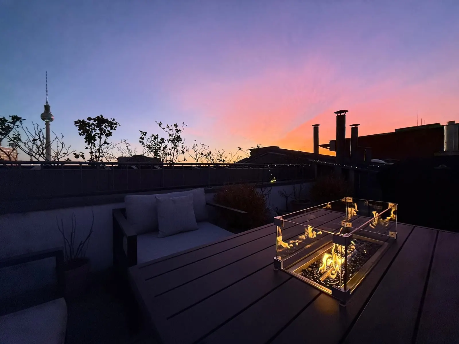 Dachterrasse mit Sofa, Tisch und Feuerschale im Abendlicht, Pflanzen am Geländer.