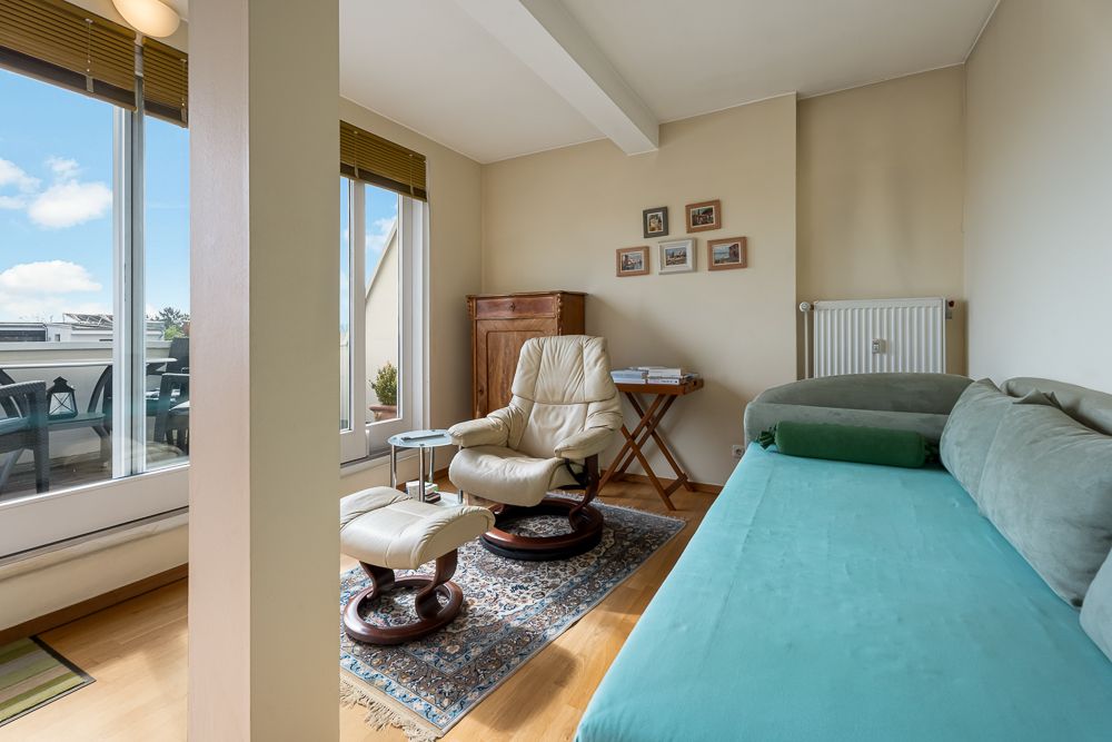 Wohnzimmer mit Sessel, Hocker, Sofa, Teppich, Holzmöbeln und großen Fenstern zum Balkon.