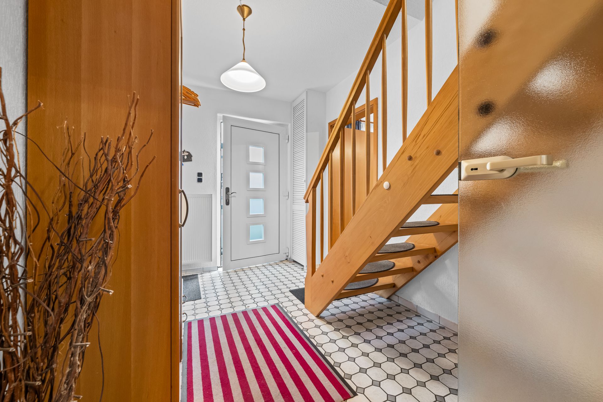 Heller Flur mit Holztreppe, weißer Eingangstür und gestreiftem Teppich auf Fliesenboden.