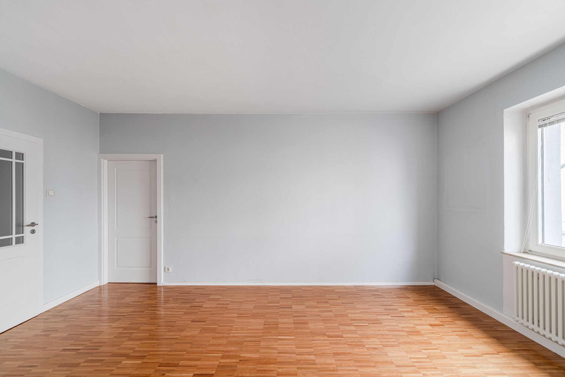 Leerer Raum mit hellem Parkettboden, weißen Türen und einem Fenster mit Heizung.
