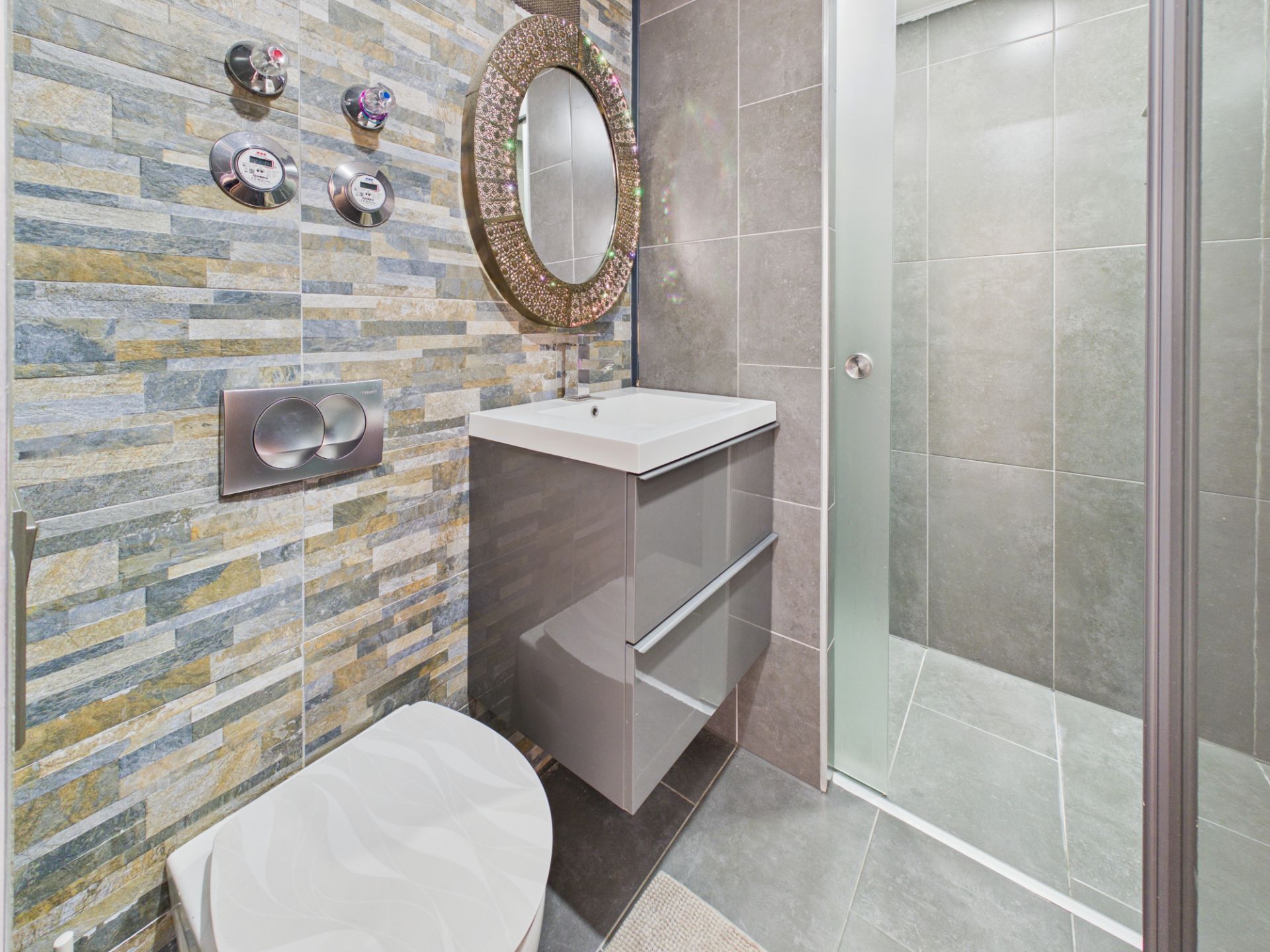 Modernes Badezimmer mit runder Spiegel, Waschbecken, Toilette und Dusche mit Glaswand.