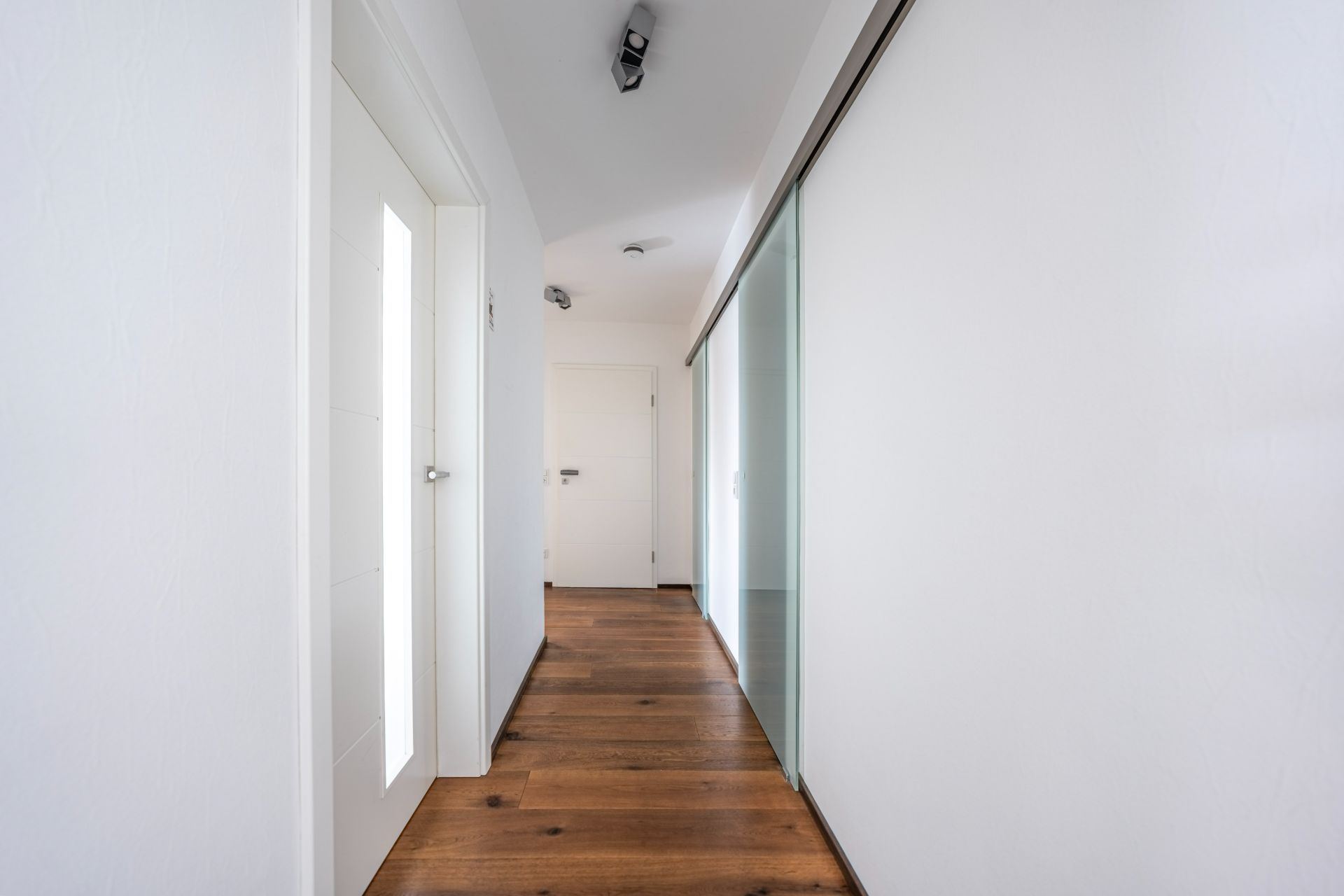 Heller Flur mit Holzboden, weißen Wänden und Türen sowie einer Glas-Schiebetür.