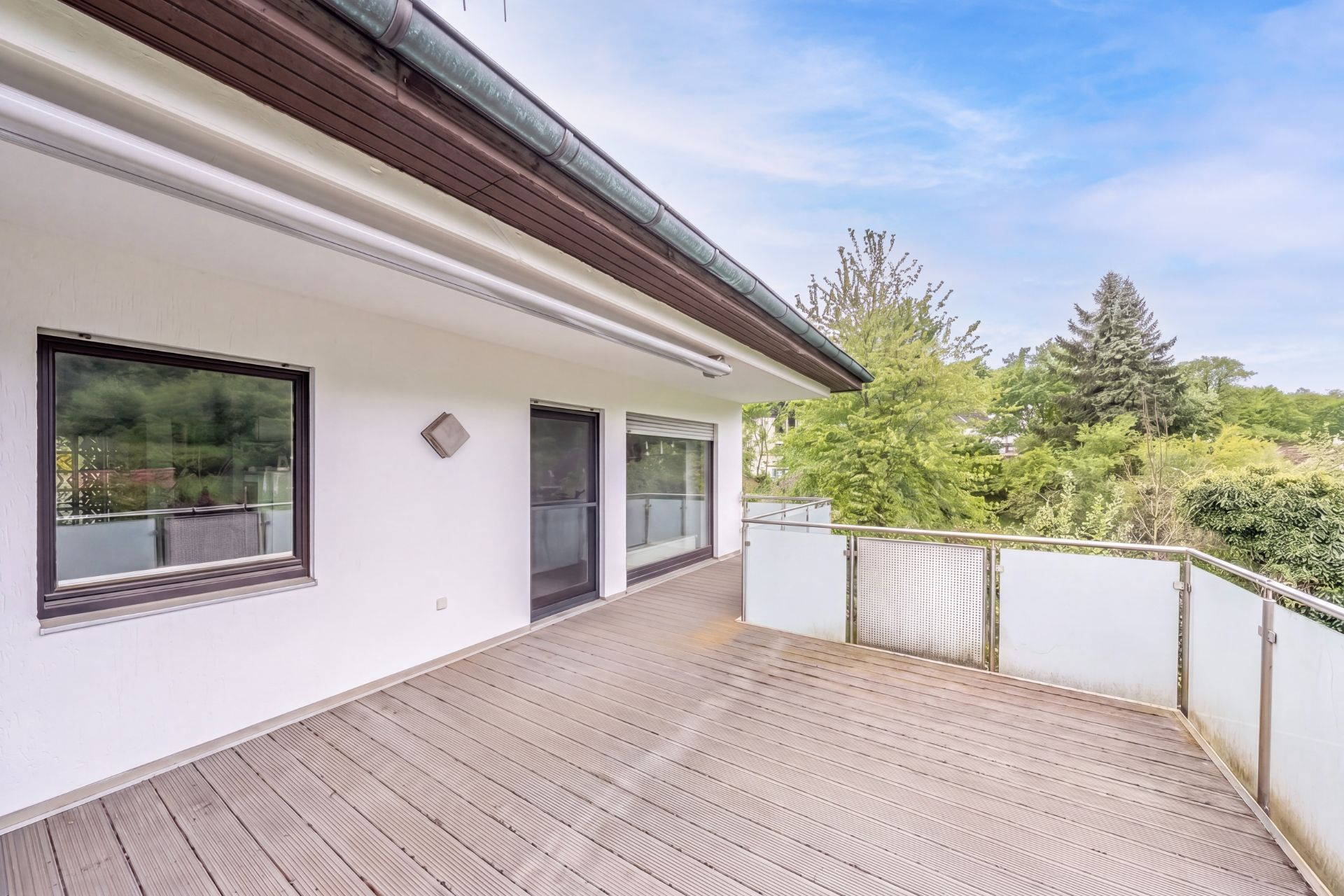 Große Terrasse mit Holzdeck, Glasgeländer und Blick auf grüne Bäume an einem Wohnhaus.