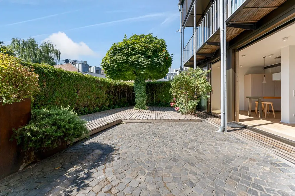 Gepflasterter Innenhof mit Baum, Hecke und offenem Zugang zur modernen Küche.