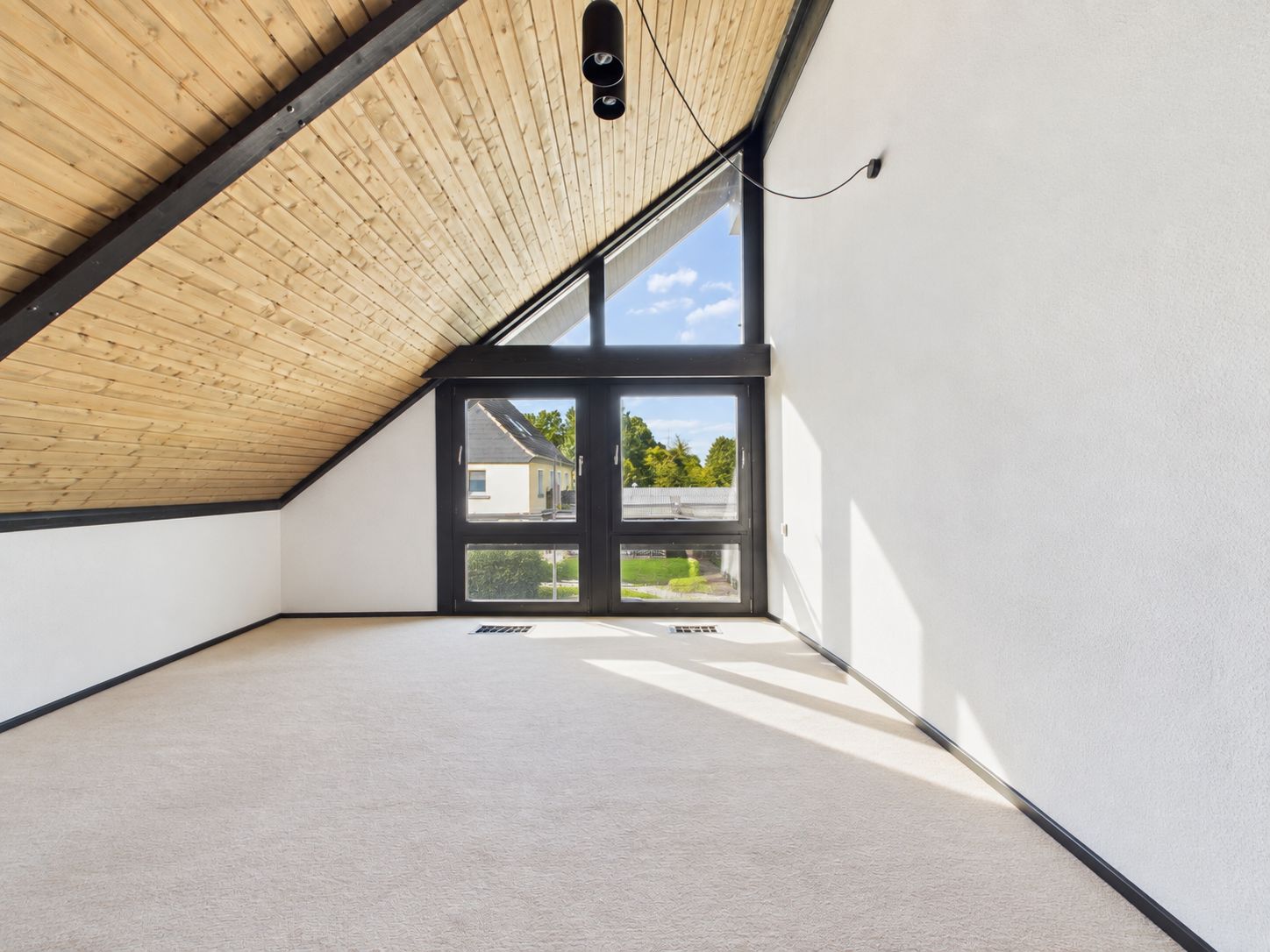 Helles Zimmer mit schräger Holzdecke, Teppichboden und großen Fenstern mit Ausblick.