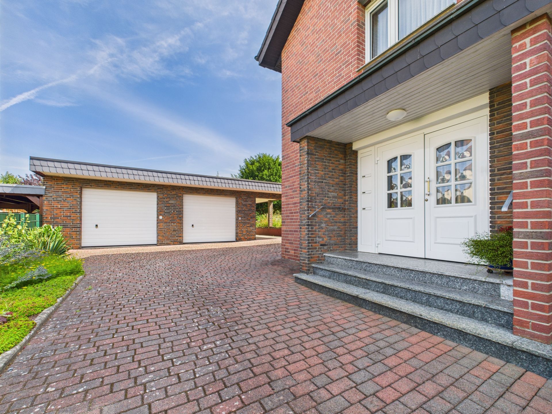 Zweiflügelige Haustür mit Treppe und gepflastertem Hof vor Doppelgarage.