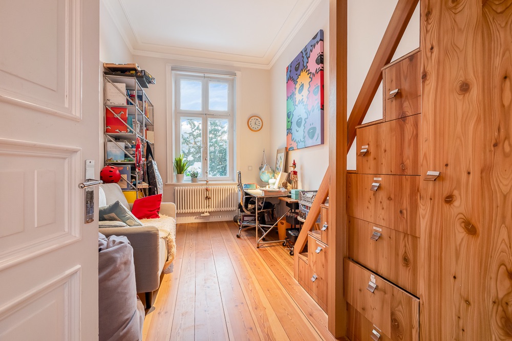 Kleines Zimmer mit Sofa, Schreibtisch, Regal, Fenster und Holztreppe mit Schubladen.