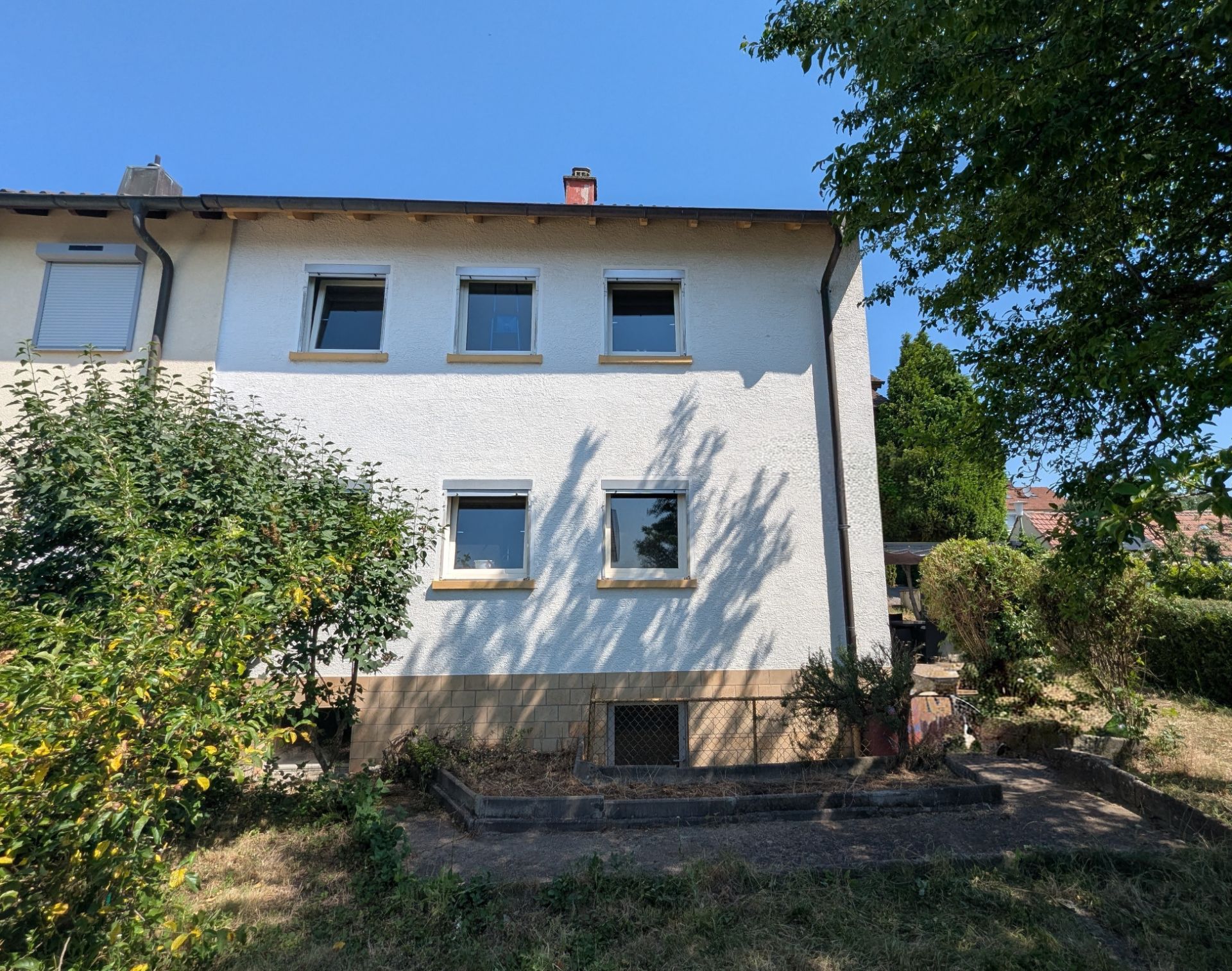 Weiße Hausfassade mit fünf Fenstern, umgeben von Garten und Bäumen, bei sonnigem Wetter.