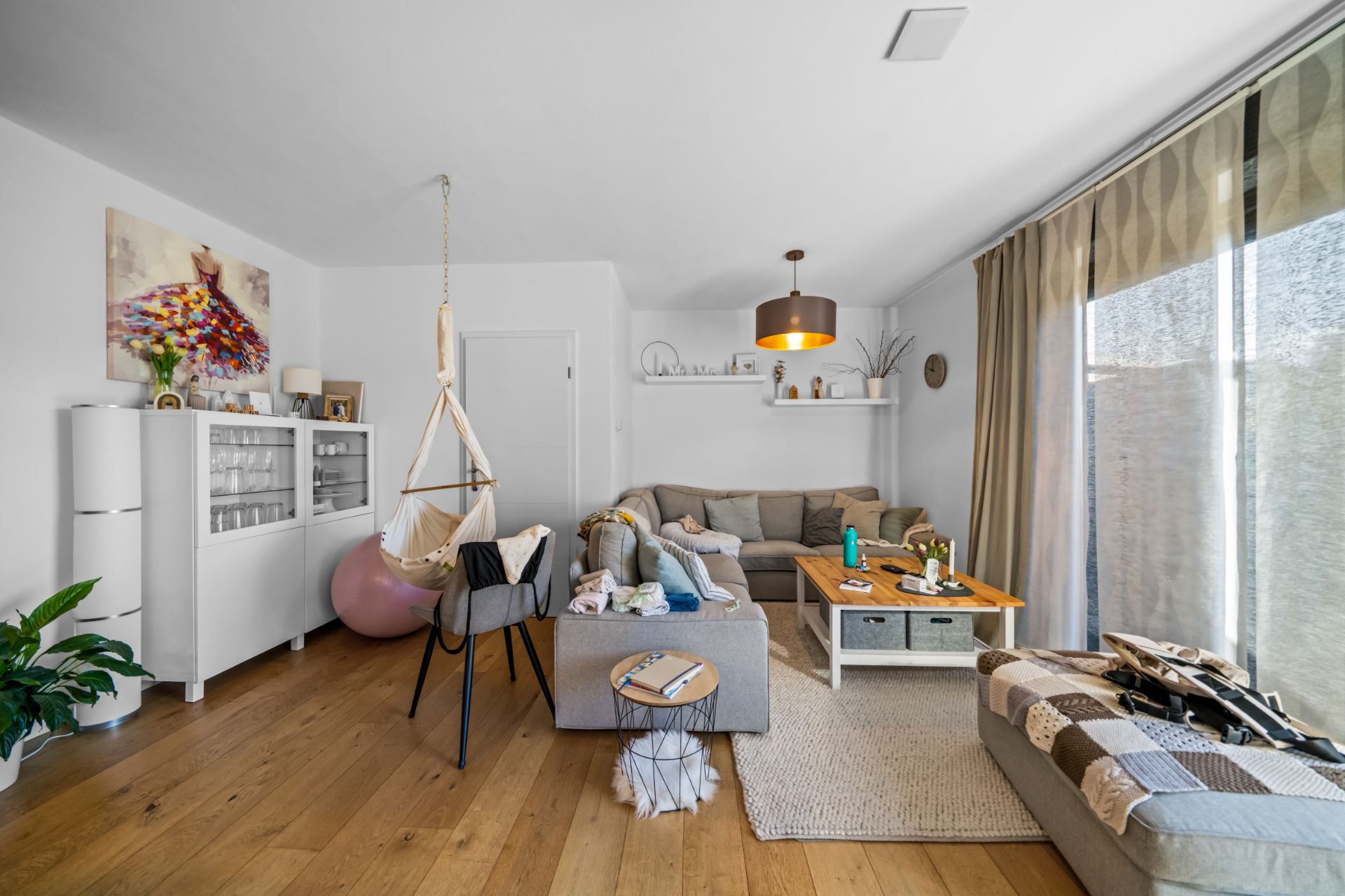 Helles Wohnzimmer mit Sofa, Sessel, Couchtisch, Hängesessel und großen Fenstern mit Vorhängen.