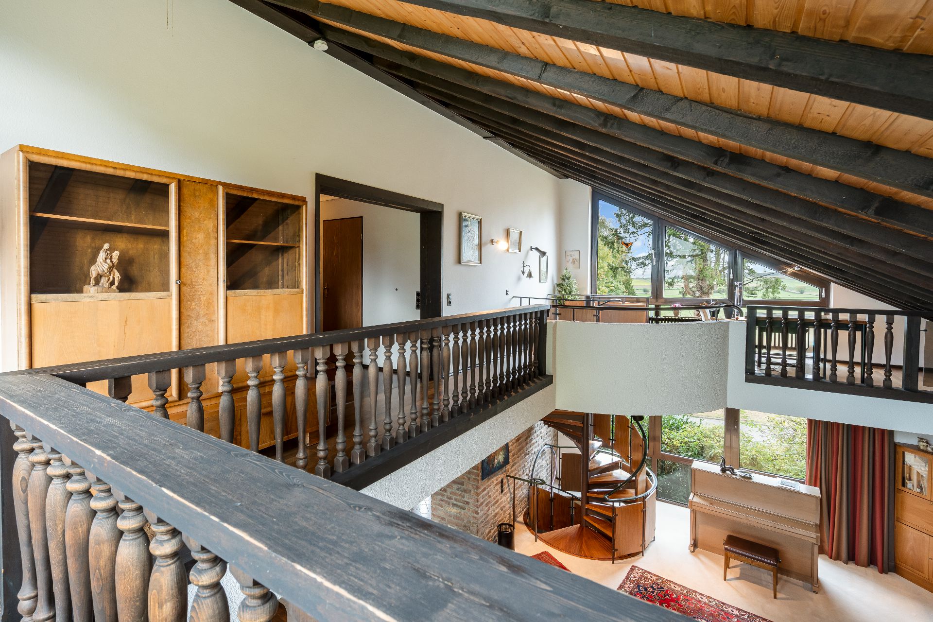 Obergeschoss mit Holzgeländer, Bücherregal und Blick auf eine Wendeltreppe und große Fenster.