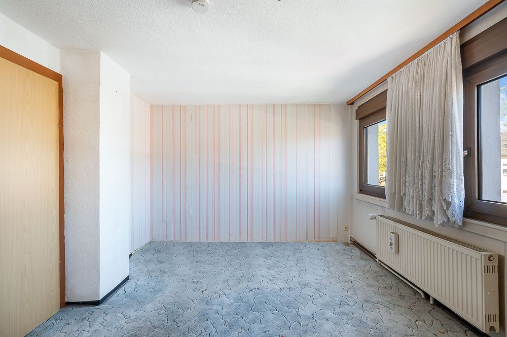 Leeres Zimmer mit gemustertem Boden, Streifentapete, zwei Fenstern und Heizkörper.