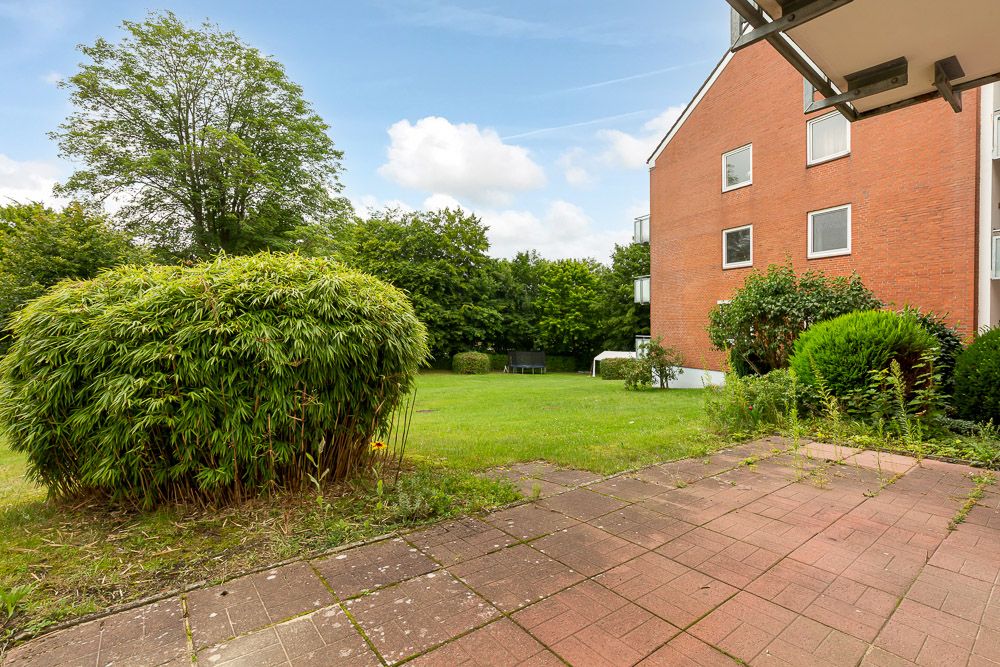 Blick von einer Terrasse auf einen Garten mit Rasen, Büschen und Bäumen neben einem Wohnhaus.