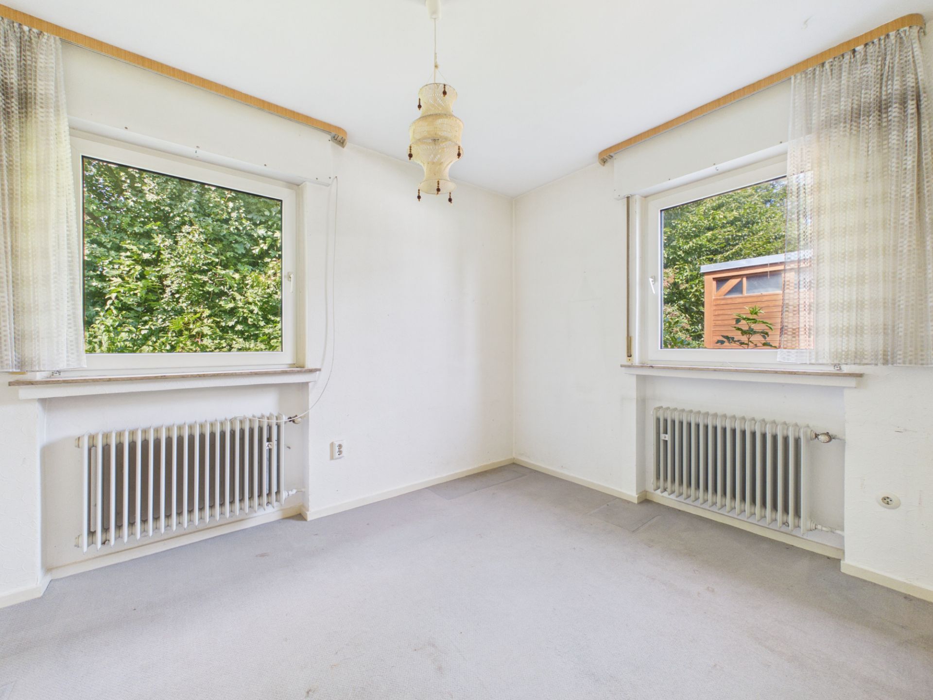 Helles Zimmer mit zwei Fenstern, Heizkörpern und hellen Vorhängen, Teppichboden.