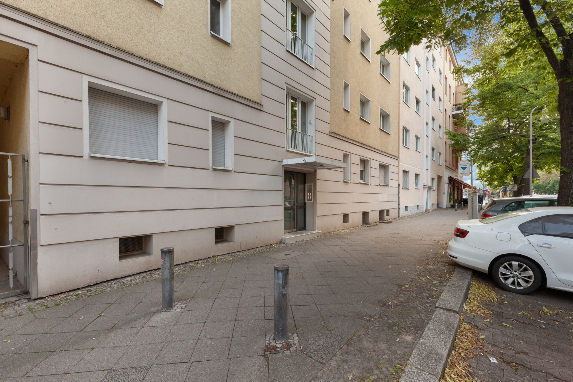 Eingang eines Mehrfamilienhauses mit Gehweg und parkenden Autos an der Straße.