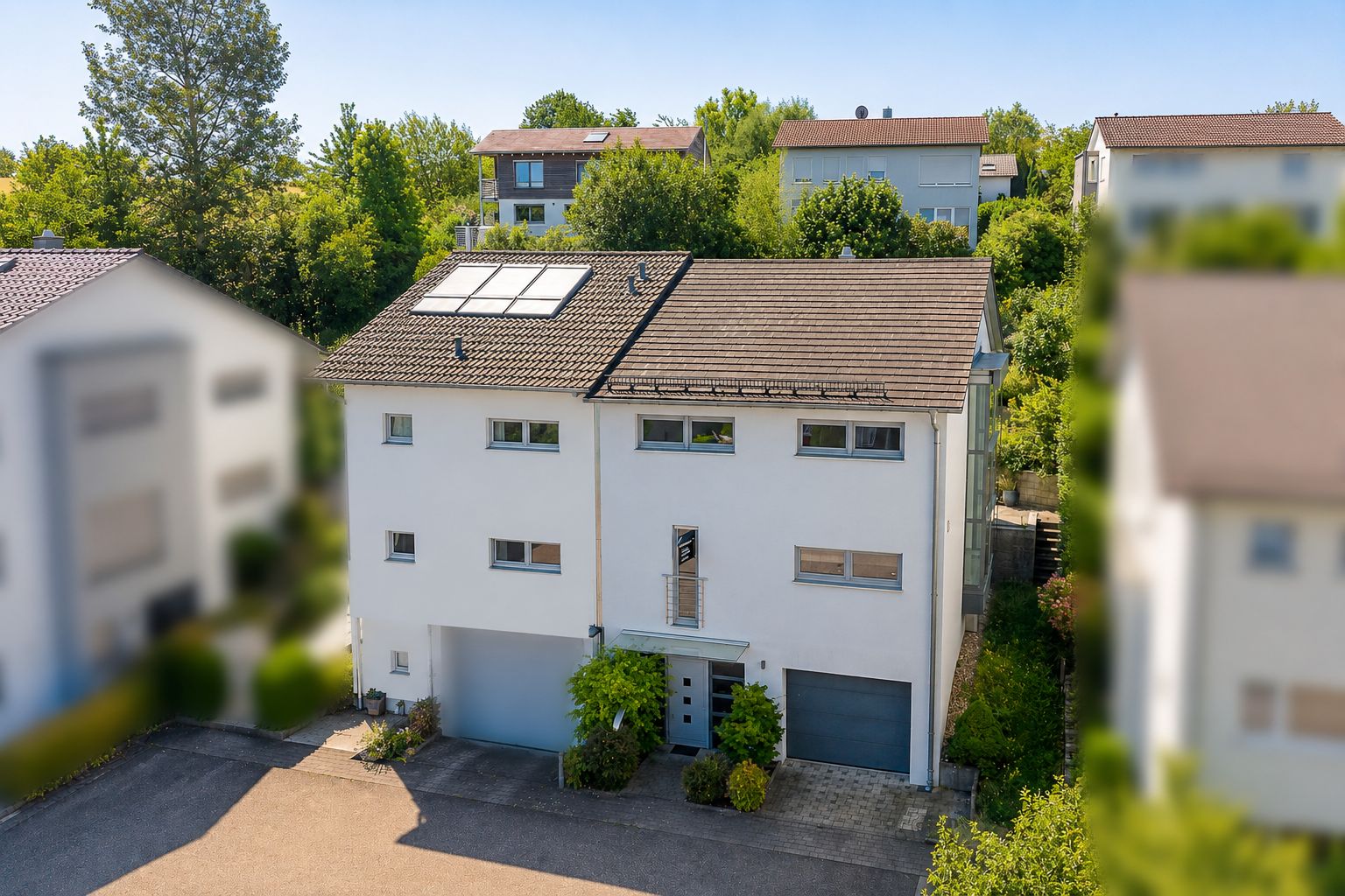 Modernes, weißes Wohnhaus mit Solarpanelen auf dem Dach und Garage, umgeben von Bäumen.