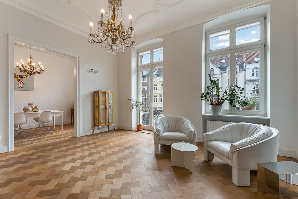 Helles Wohnzimmer mit zwei weißen Sesseln, Holzboden, Kronleuchter und großen Fenstern.