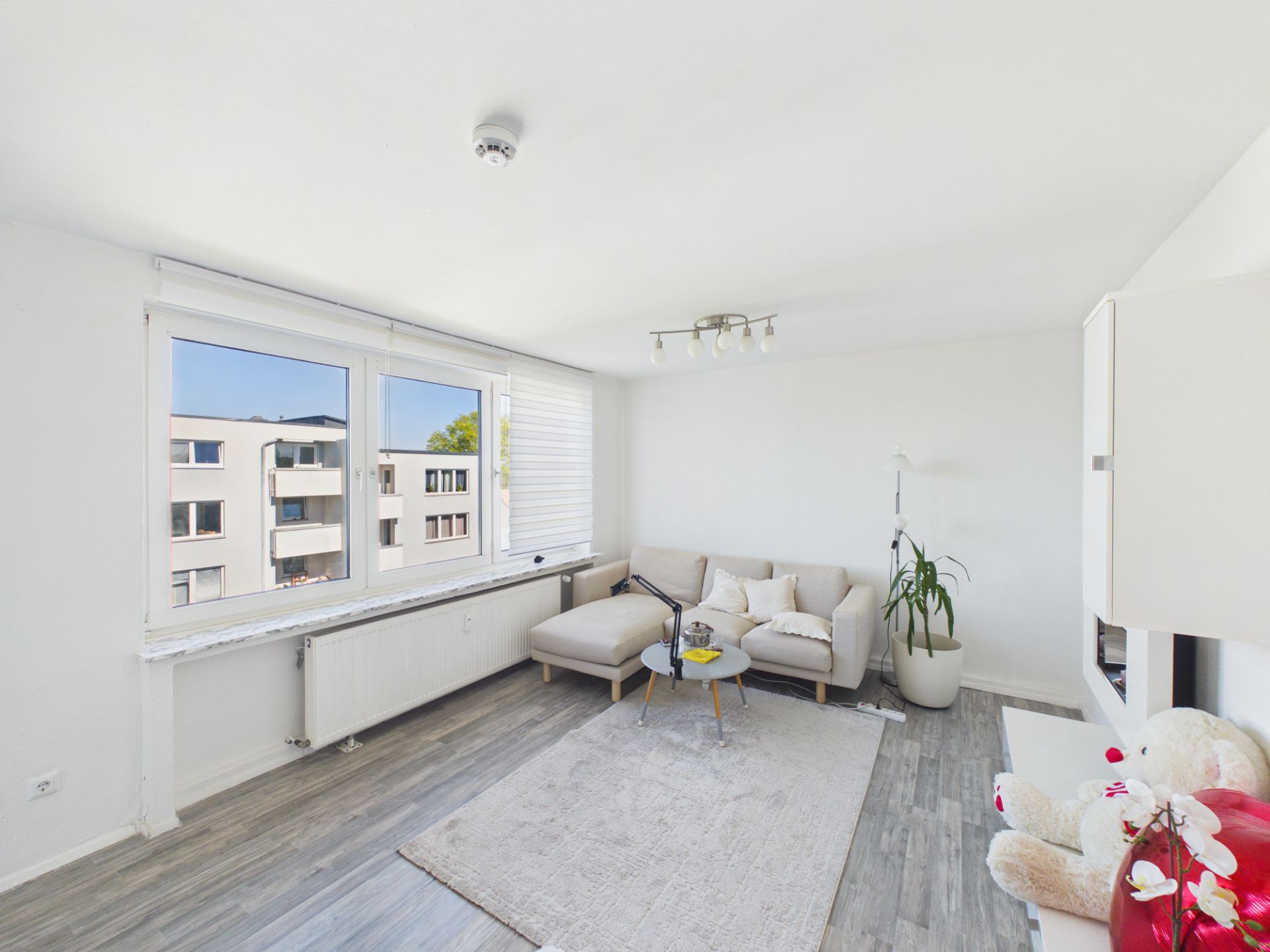 Helles Wohnzimmer mit Ecksofa, Teppich, Pflanze und großem Fenster.