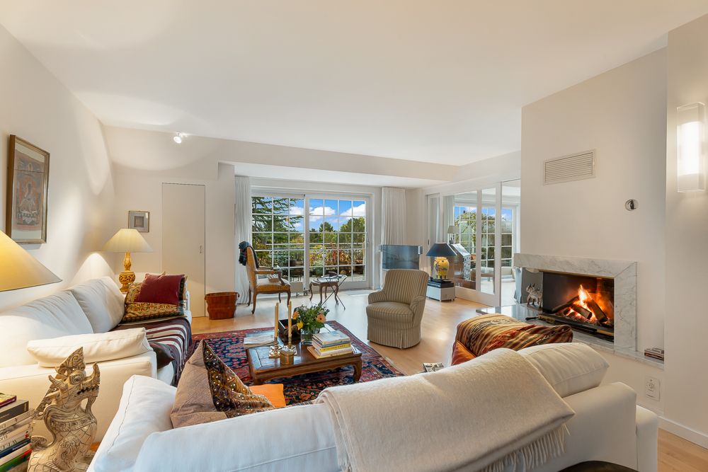 Helles Wohnzimmer mit Kamin, Sofas, Teppich und großen Fenstern mit Blick auf den Garten.