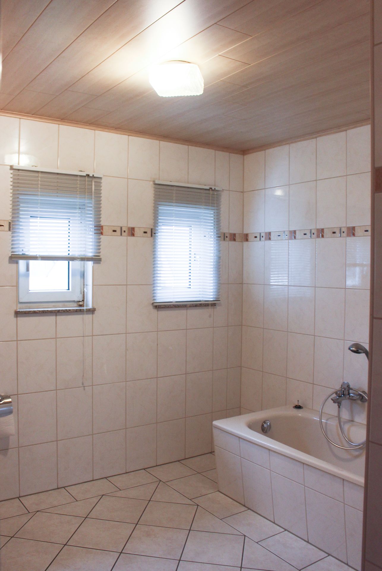 Helles Badezimmer mit Badewanne, zwei Fenstern mit Jalousien und weißen Fliesen.