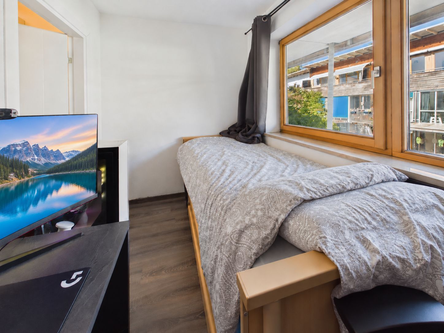 Kleines Schlafzimmer mit Einzelbett, großem Fenster und Schreibtisch mit Monitor.