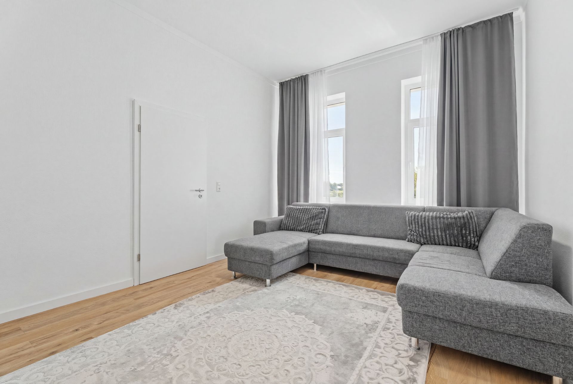 Helles Wohnzimmer mit grauem Ecksofa, Teppich, Holzfußboden und zwei Fenstern mit Gardinen.