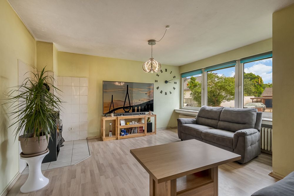 Wohnzimmer mit grauem Sofa, großem Fernseher, Holztisch, Pflanze und großen Fenstern mit Blick nach draußen.