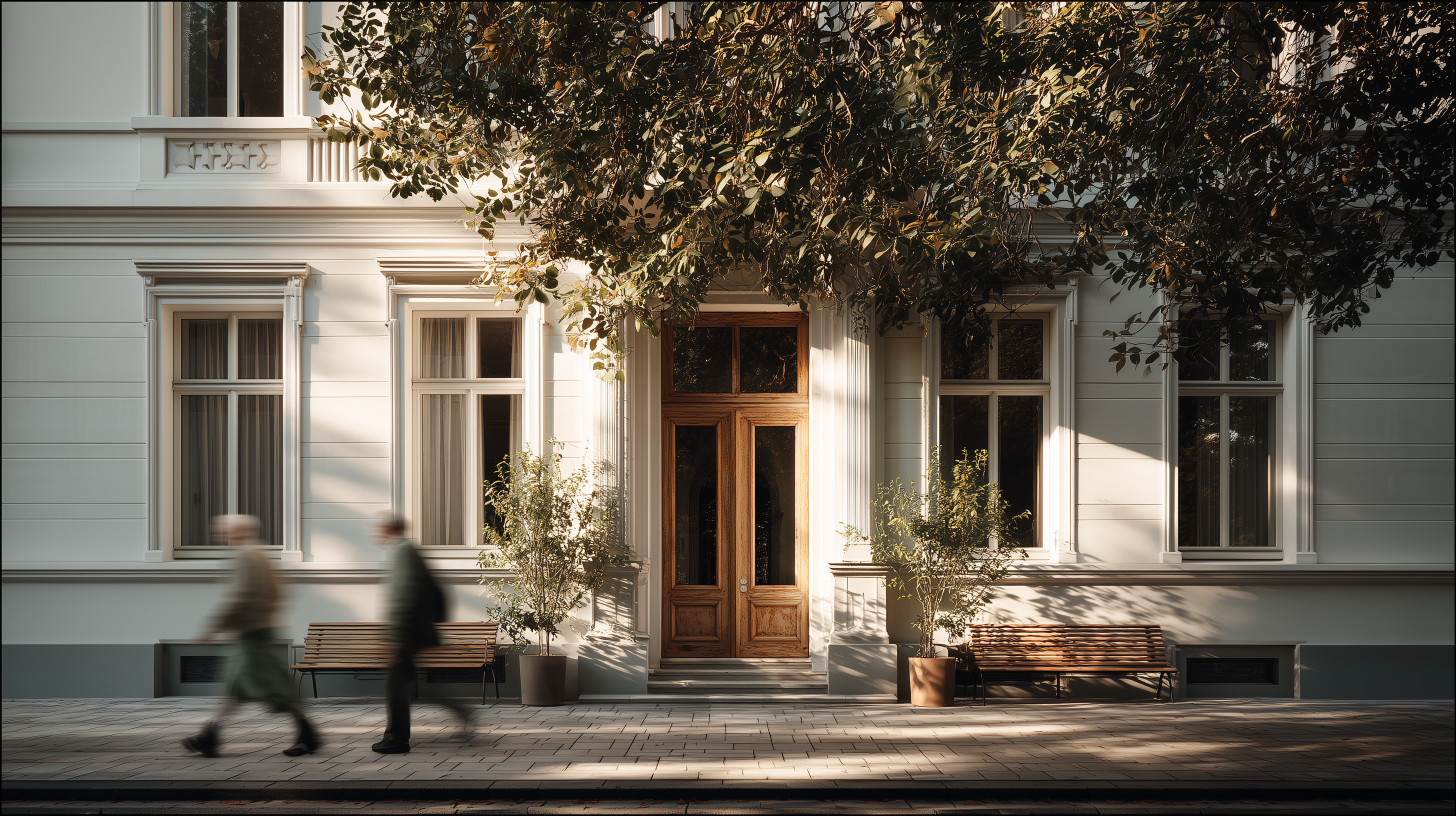 Weiße Hausfassade mit Holztür, zwei großen Fenstern, Bänken, Pflanzen und zwei vorbeigehenden Personen.