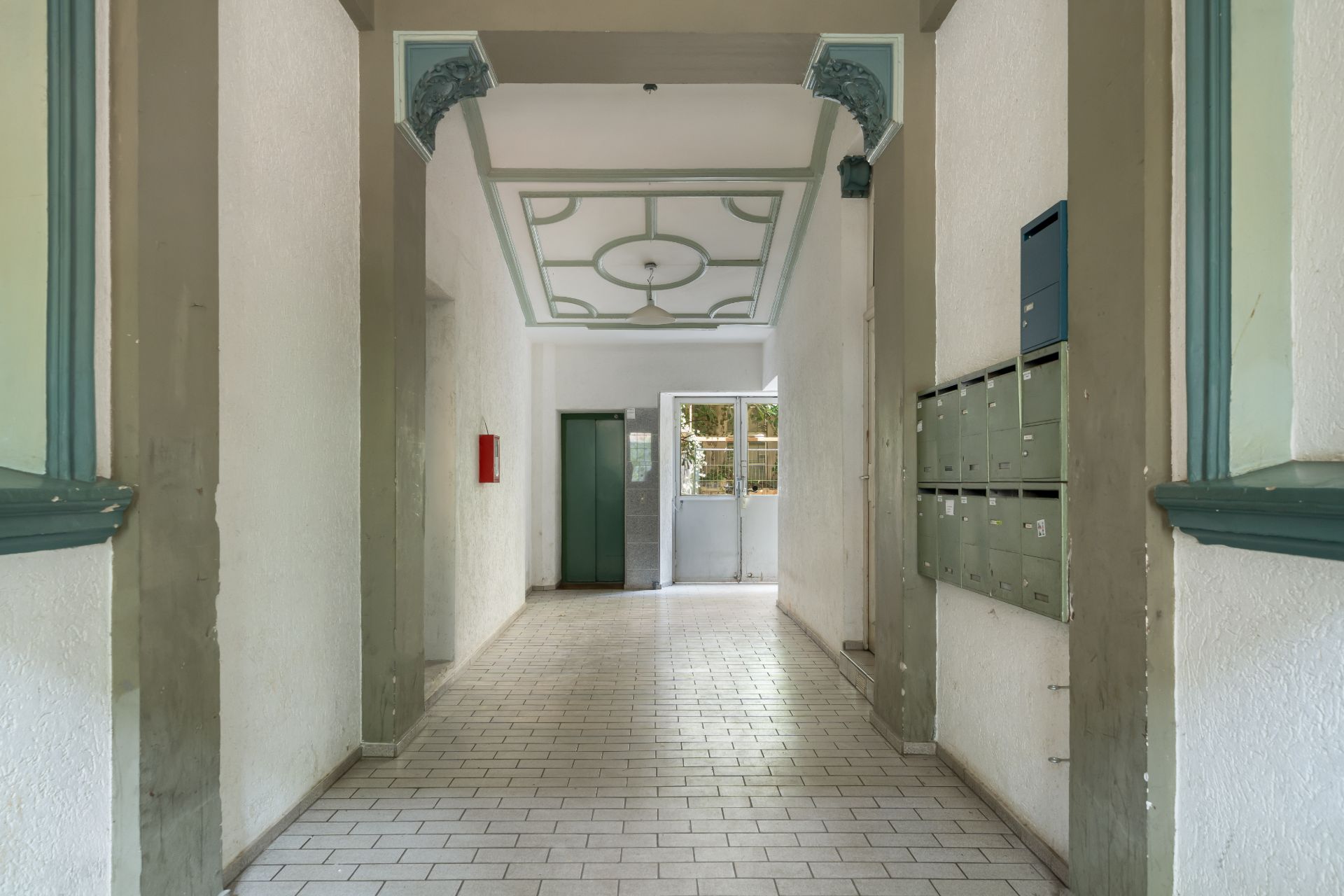 Heller Flur mit Briefkästen, Aufzug und Tür am Ende, weiße Wände und Bodenfliesen.