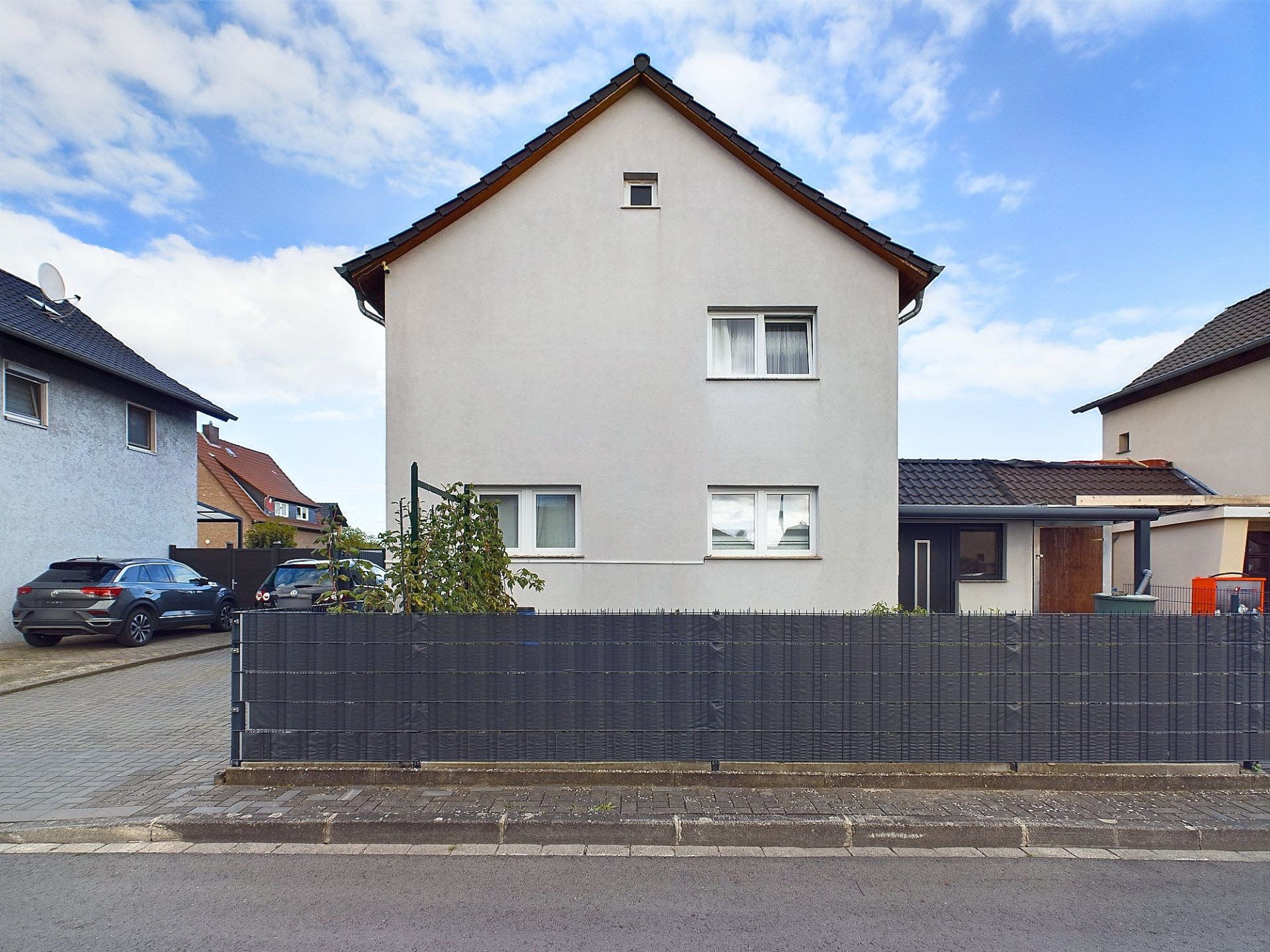 Zweistöckiges Wohnhaus mit hellem Putz, schwarzen Dachziegeln und Zaun im Vordergrund.
