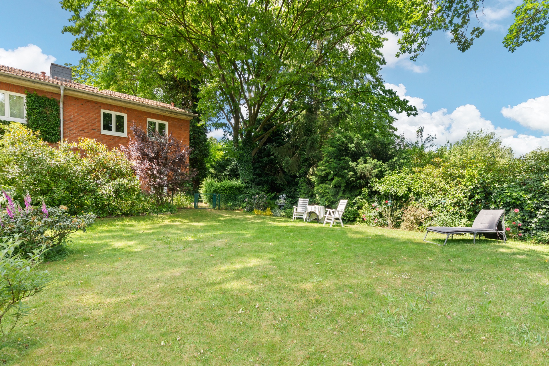 Großer grüner Garten mit Liege, Sitzgruppe und Bäumen neben einem roten Wohnhaus.