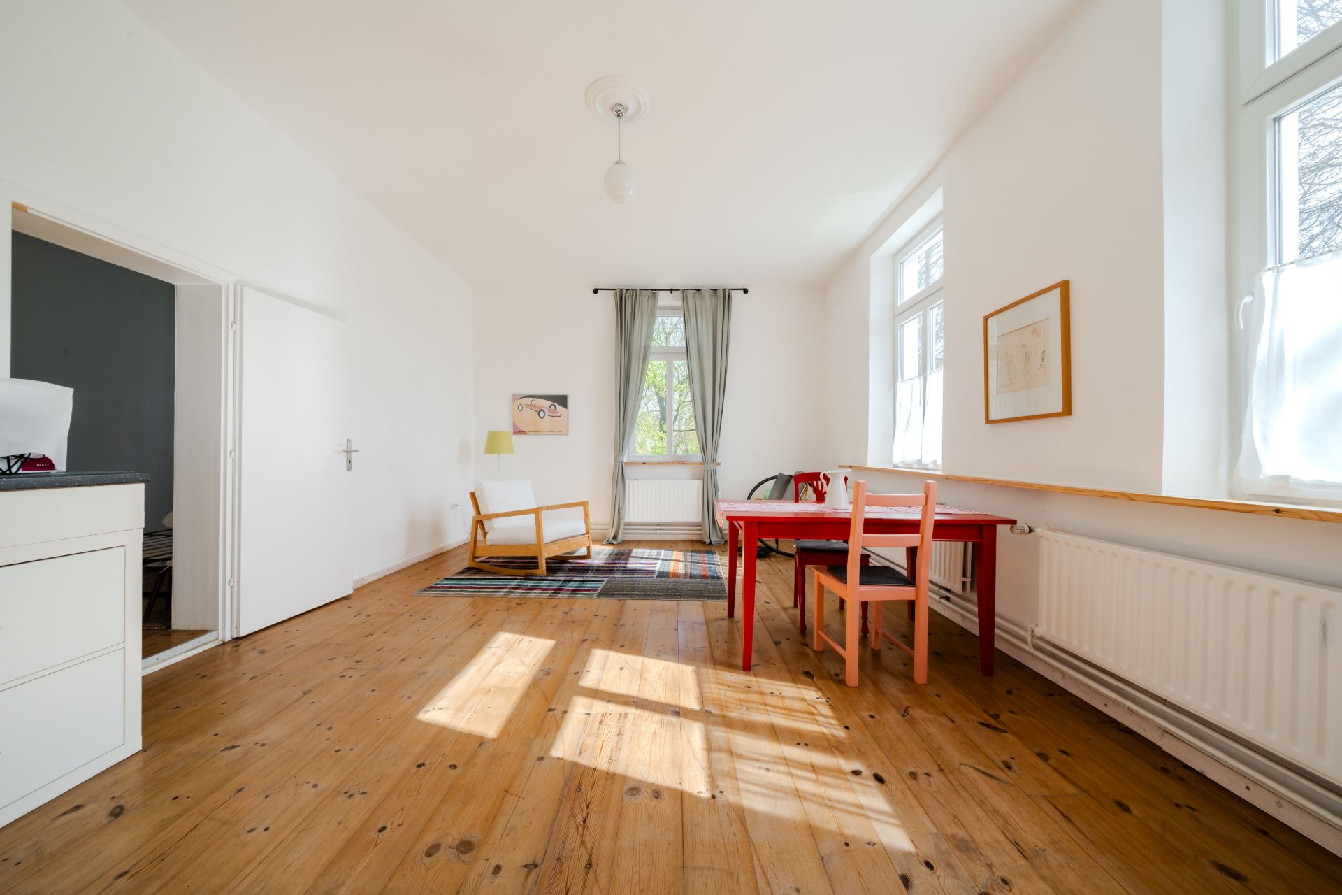 Helles Zimmer mit Holzboden, Tisch, zwei Stühlen, Sessel, Fenster und Vorhang.