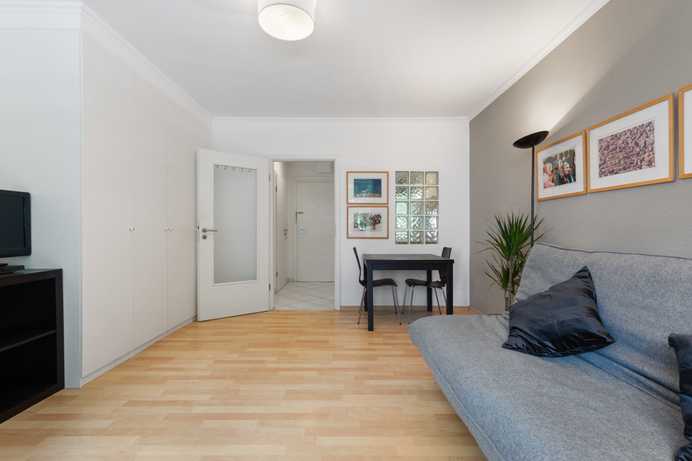 Wohnzimmer mit grauem Sofa, kleinem Esstisch, weißen Schränken und Wandbildern.