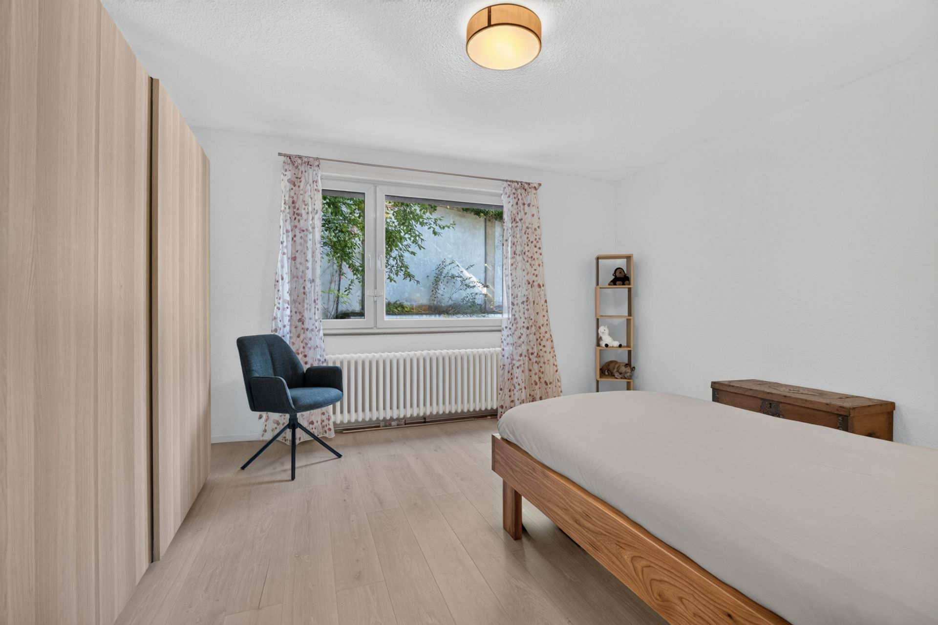 Helles Schlafzimmer mit Bett, Stuhl, Kleiderschrank, Regal und Fenster mit Blick nach draußen.