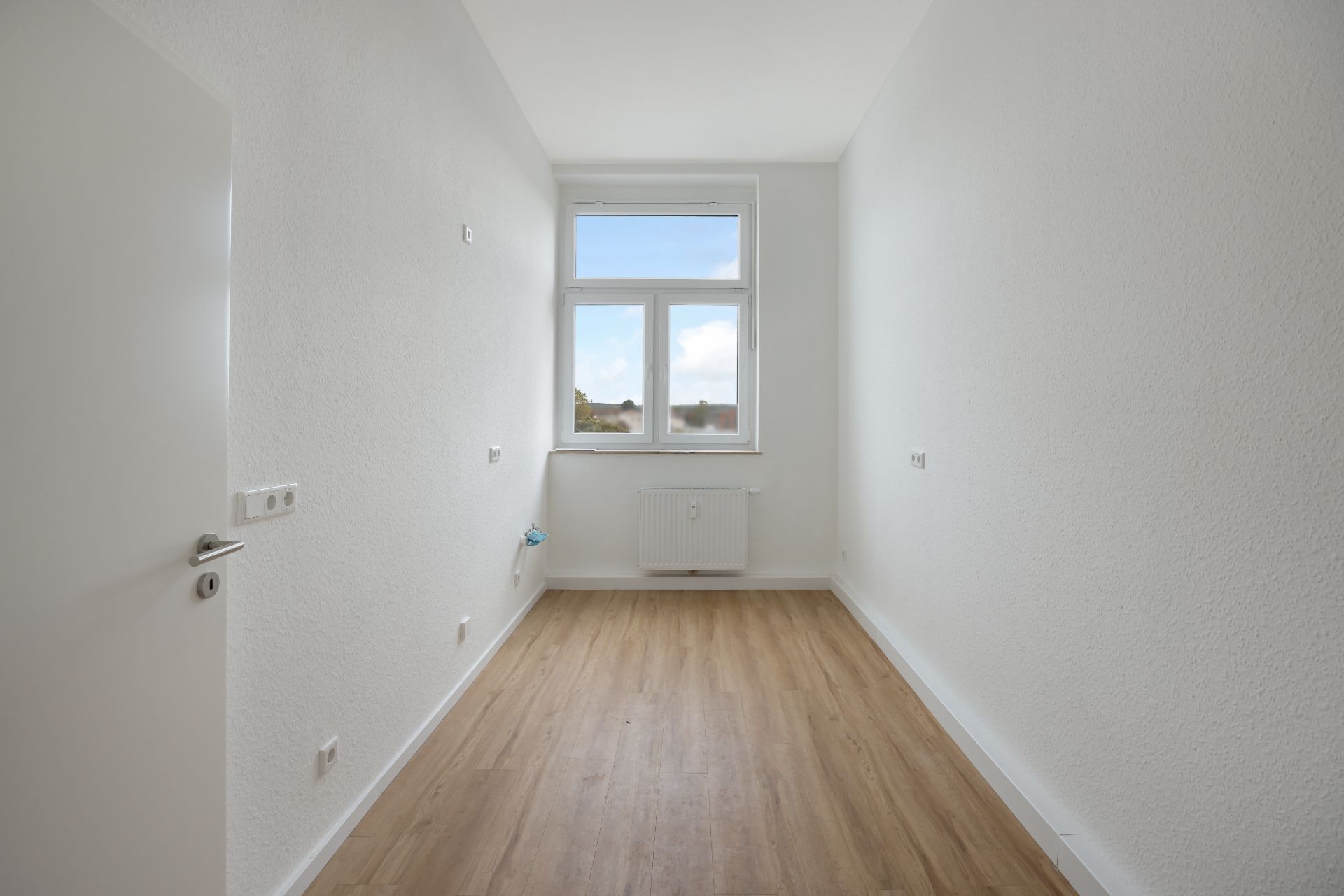 Schmaler, leerer Raum mit weiß gestrichenen Wänden, Holzfußboden und großem Fenster.