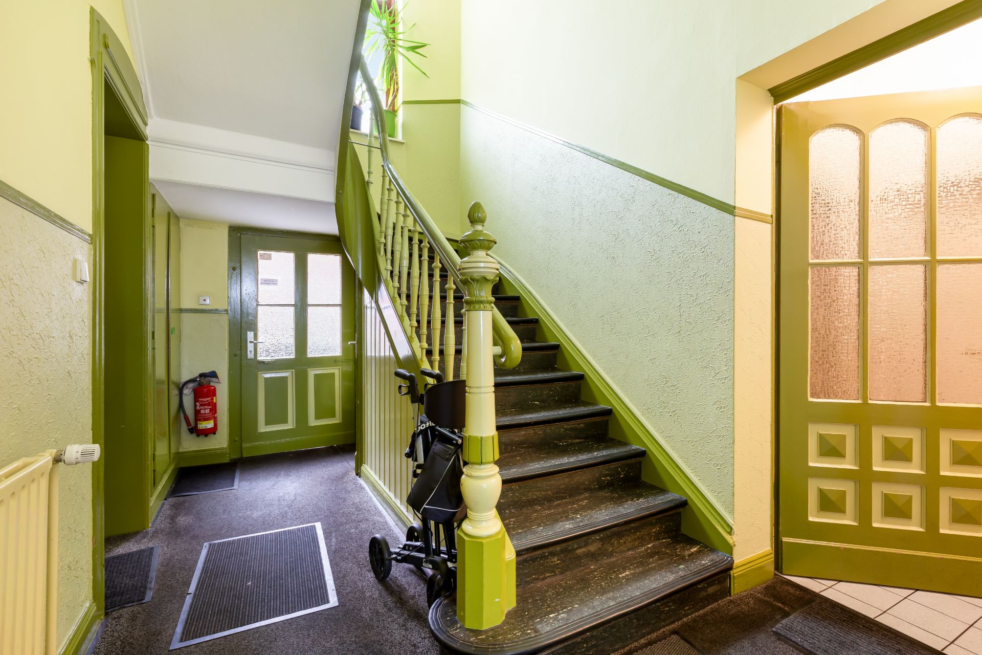 Heller Hausflur mit grüner Treppe, Kinderwagen und Glastür.