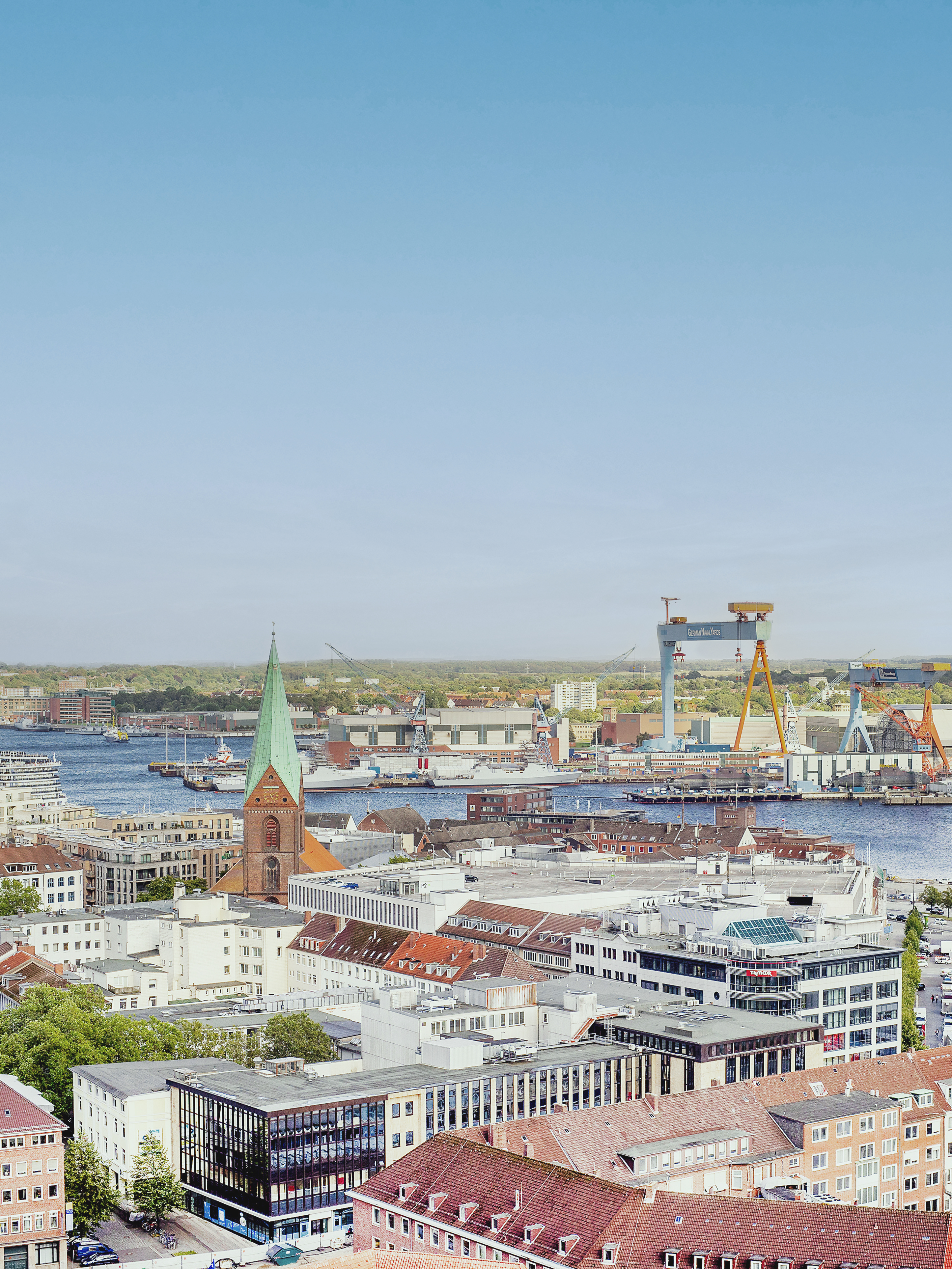 Blick auf eine Stadt mit Kirche, Häusern, Hafen und Kränen am Wasser.