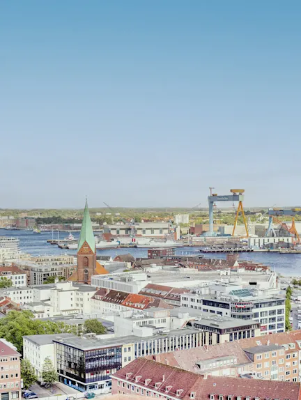 Blick auf eine Stadt mit Kirche, Häusern, Hafen und Kränen am Wasser.