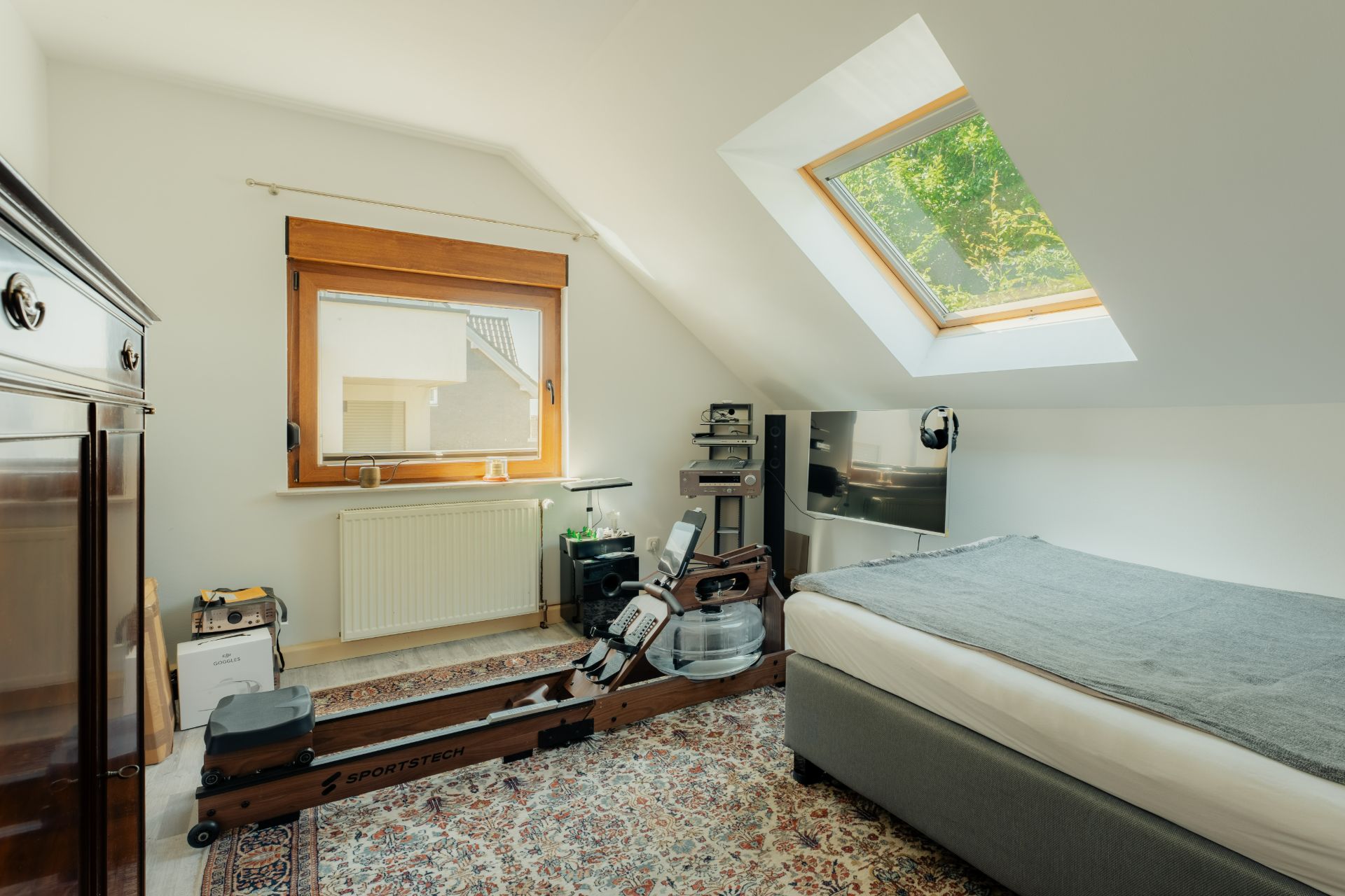 Helles Schlafzimmer mit Rudergerät, Bett, Teppich und Dachfenster.