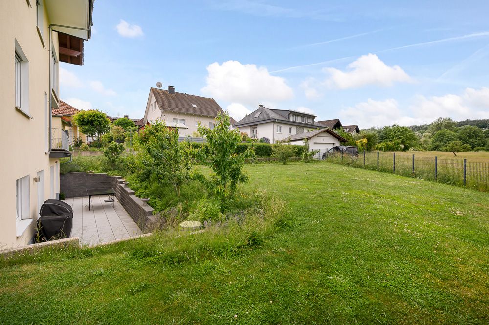 Grüner Garten mit Rasenfläche, Terrasse und benachbarten Wohnhäusern im Hintergrund.