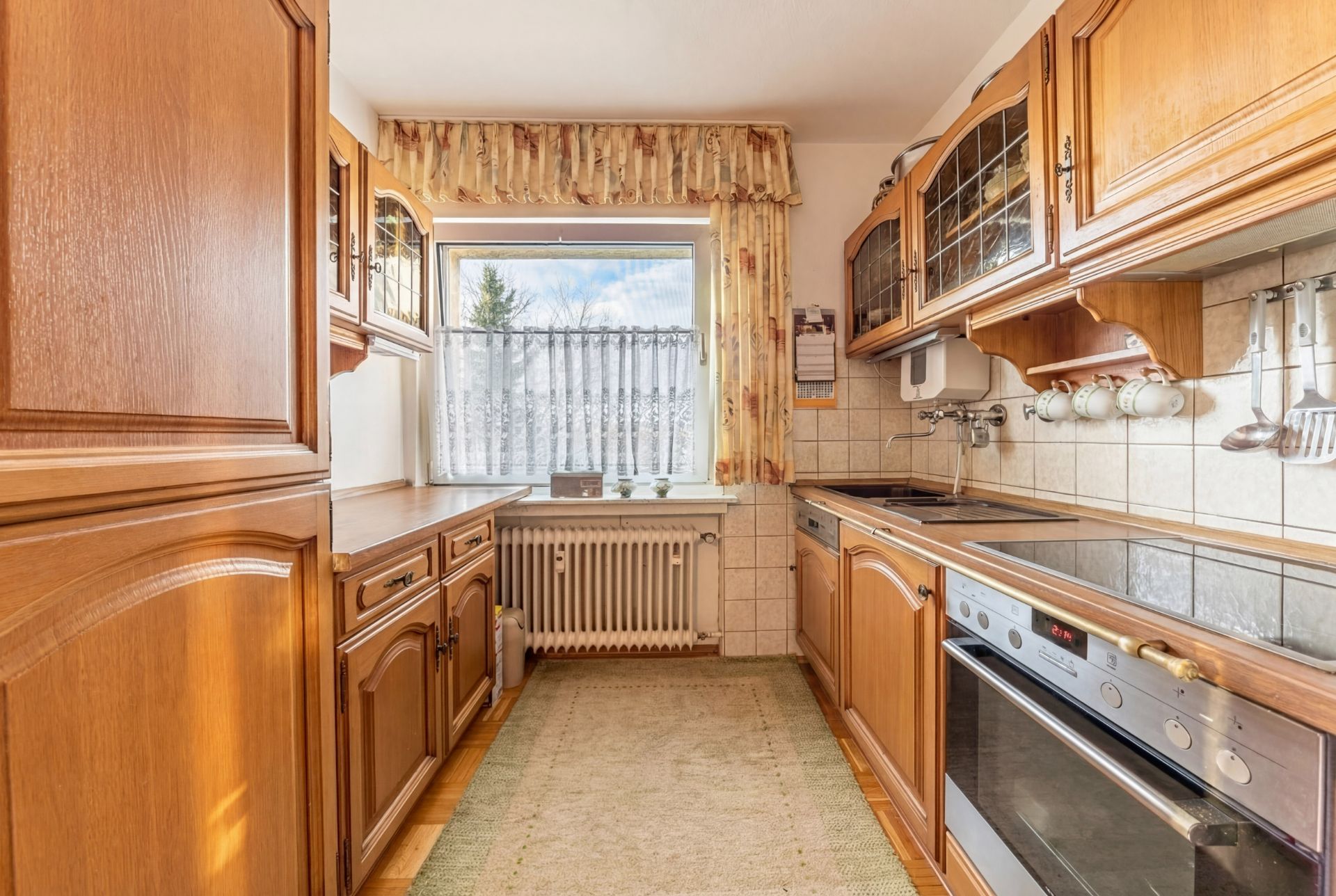 Helle Küche mit Holzschränken, Herd, Ofen und Fenster mit Vorhängen.