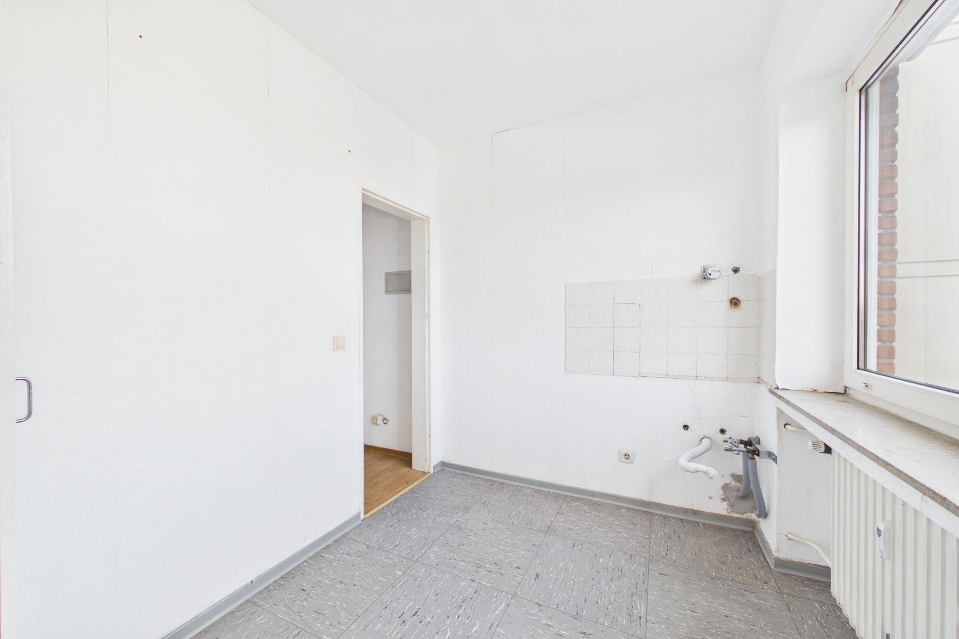 Leerer, heller Raum mit Fliesenboden, Wasseranschlüssen und großem Fenster.