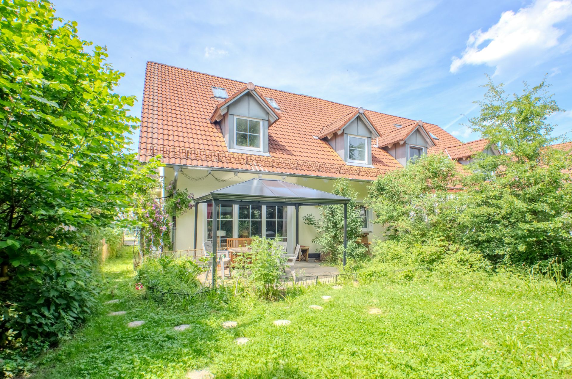 Zweistöckiges Haus mit rotem Dach, Wintergarten und Garten mit grünen Bäumen und Rasen.