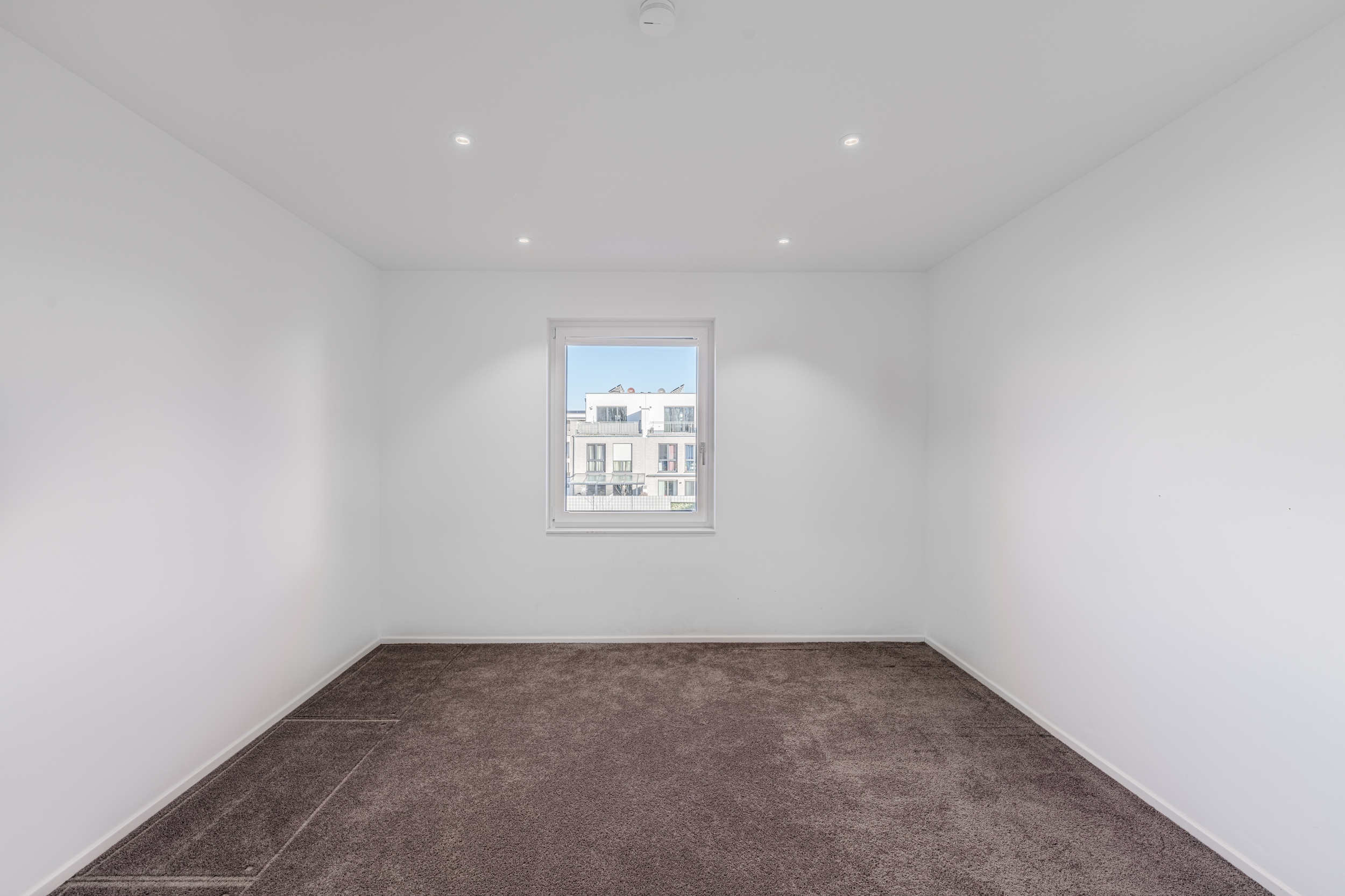 Leerer Raum mit weiß gestrichenen Wänden, braunem Teppichboden und einem Fenster.