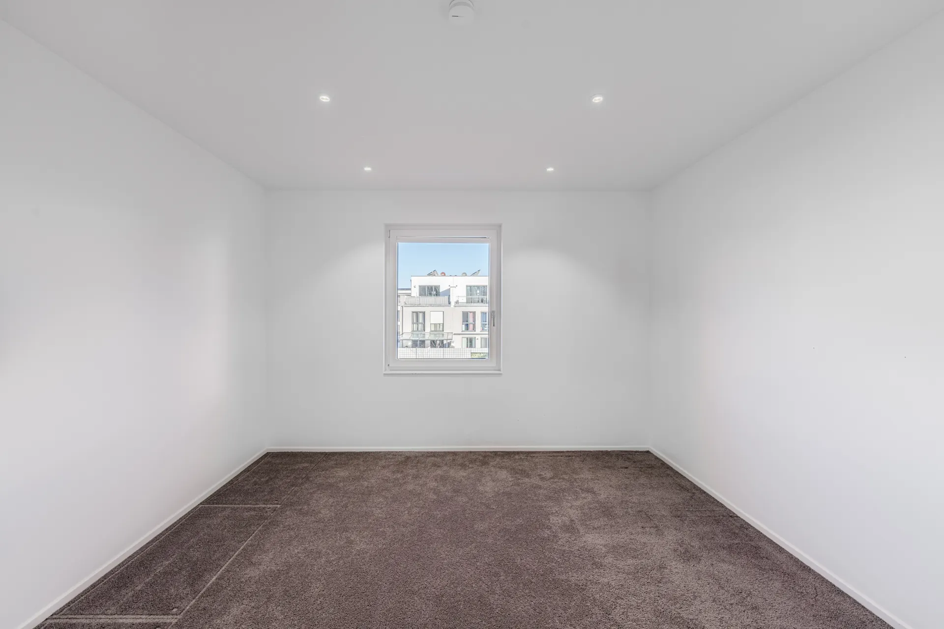 Leerer Raum mit weiß gestrichenen Wänden, braunem Teppichboden und einem Fenster.