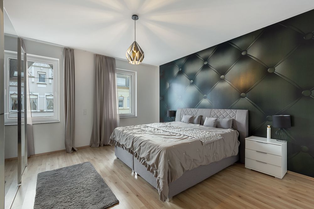 Modernes Schlafzimmer mit großem Bett, zwei Fenstern und dekorativer Wand hinter dem Bett.