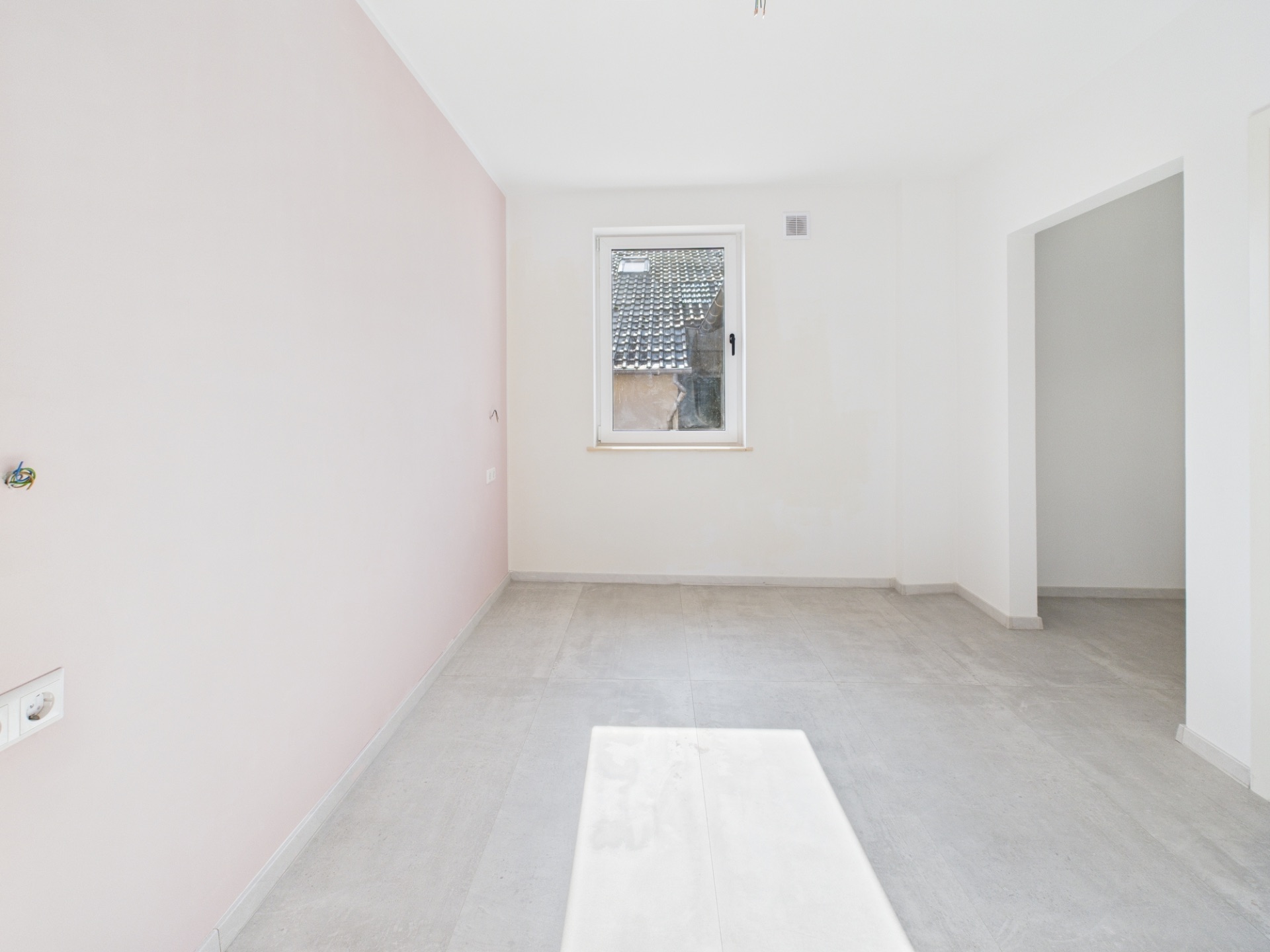 Heller, leerer Raum mit Fenster, rosa Wand links und Durchgang rechts.
