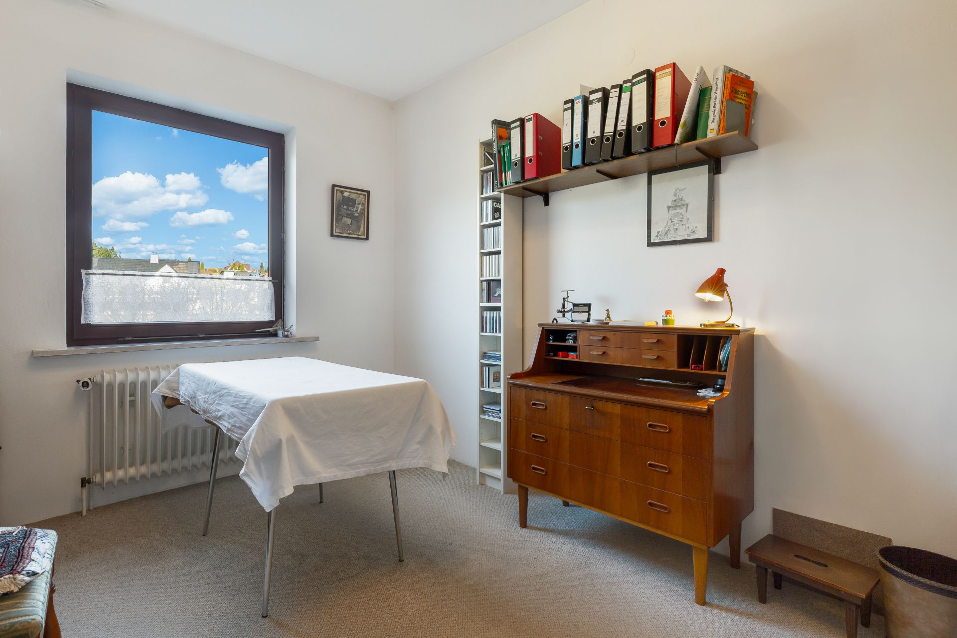 Helles Zimmer mit Schreibtisch, Bücherregal, Fenster und Tisch mit weißem Tuch.