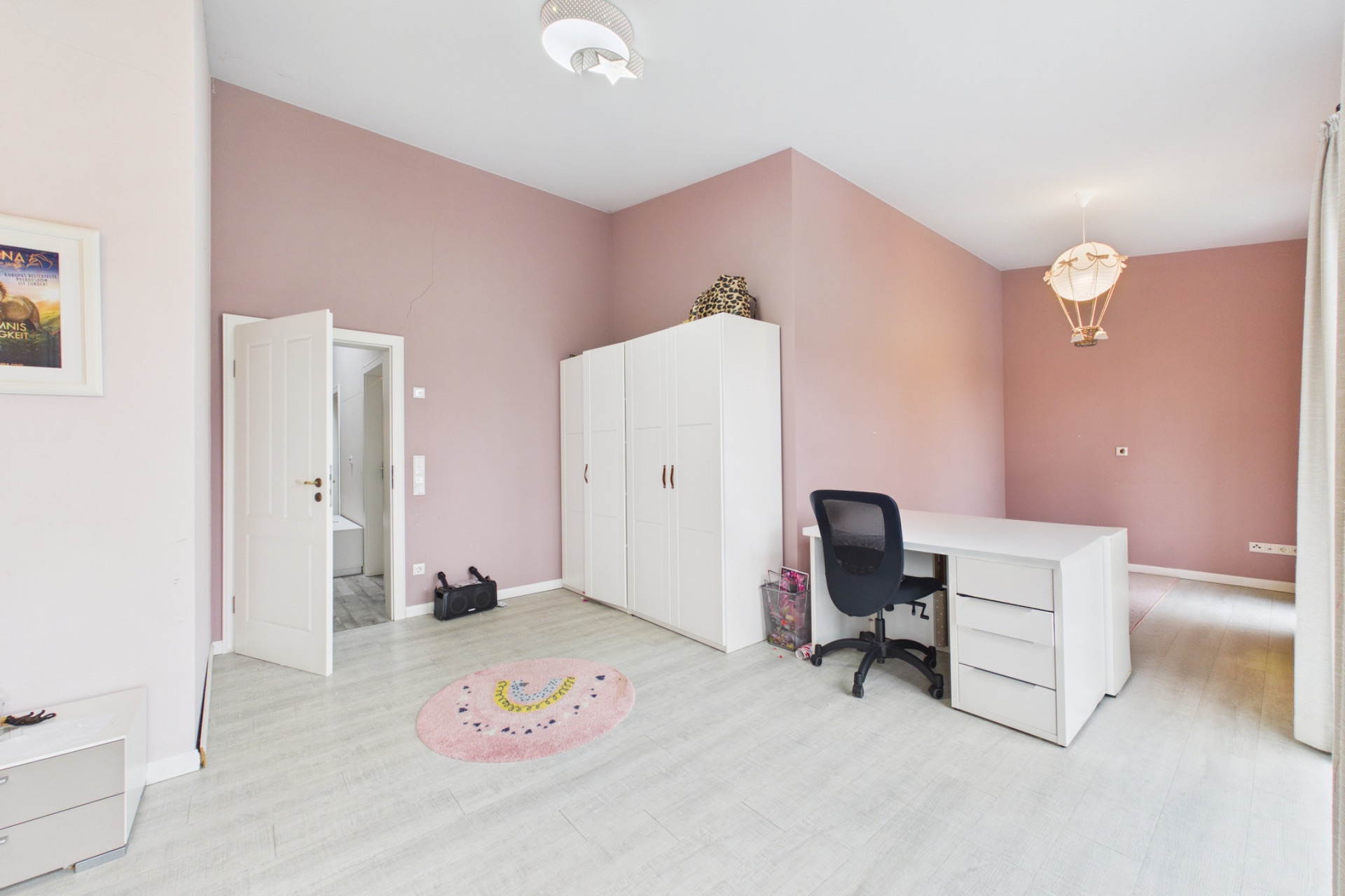Helles Zimmer mit rosa Wänden, Schreibtisch, Bürostuhl, Kleiderschrank und rundem Teppich.