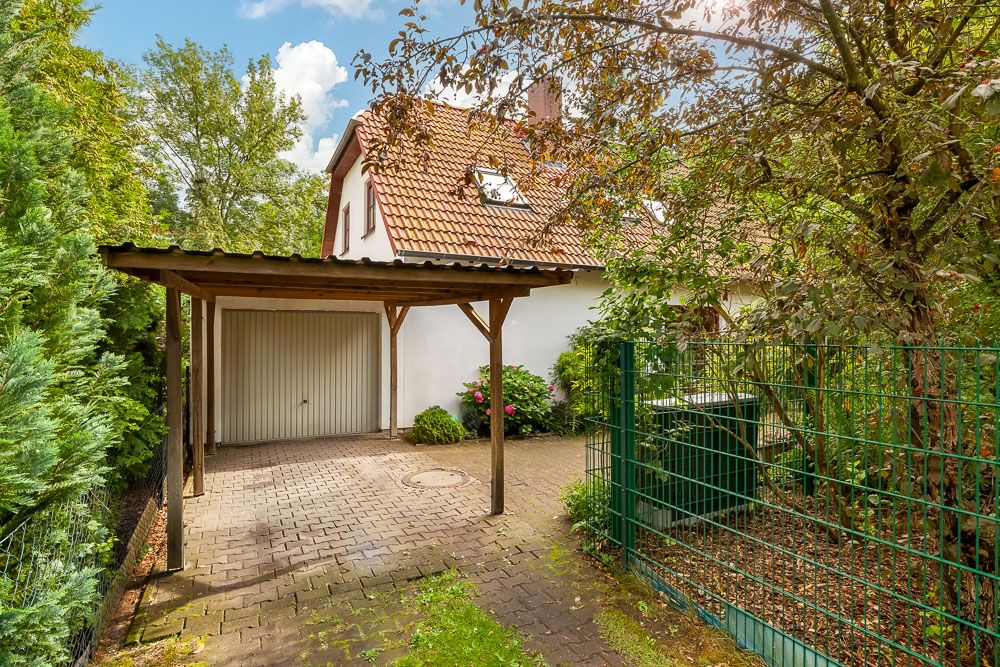 Haus mit rotem Dach, Carport, Garage und Garten, umgeben von Bäumen und Zaun.